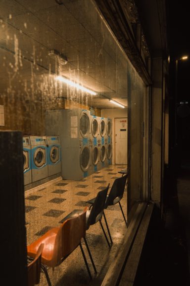 A dimly lit laundrette with washing machines and empty chairs visible through a window.