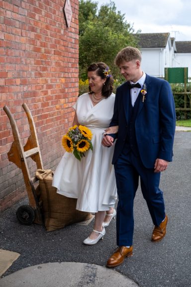 Bride in a white dress and groom in a blue suit walking together, holding sunflowers.
